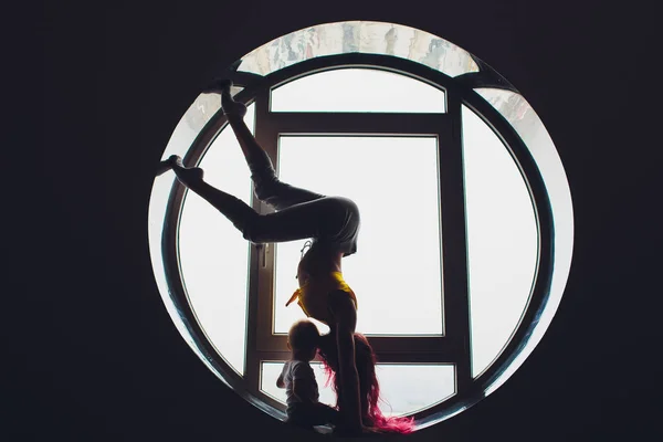 La silueta oscura de una hermosa mujer con niño en el fondo de la ventana redonda. yoga, acrobacias . — Foto de Stock