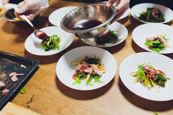 Un chef masculino decora un plato de ensalada en una cocina de restaurante en un delantal negro. primer plano de una mano con pinzas . — Foto de Stock
