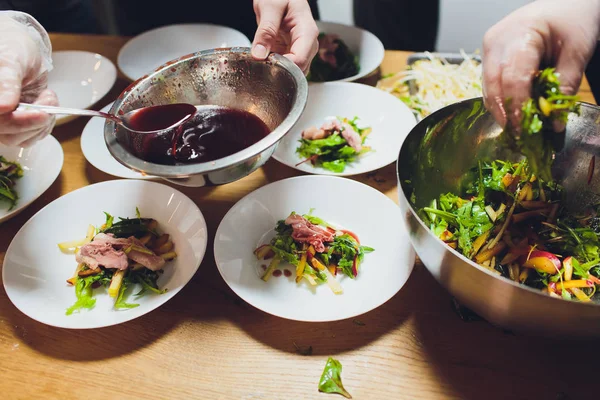 Un chef masculino decora un plato de ensalada en una cocina de restaurante en un delantal negro. primer plano de una mano con pinzas . — Foto de Stock