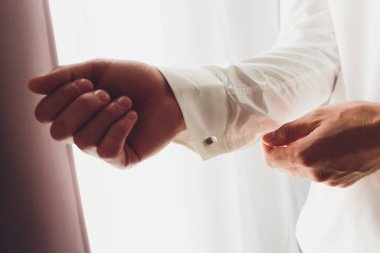 Adam gömleğin kelepçelerini bağlatıyor. Damadın sabahı. Düğün damadıbeyaz gömleğini ilikleyerek. Erkekler beyaz bir gömlek, kol düğmeli kollu gömlek bir arka plan üzerinde eller. Close.