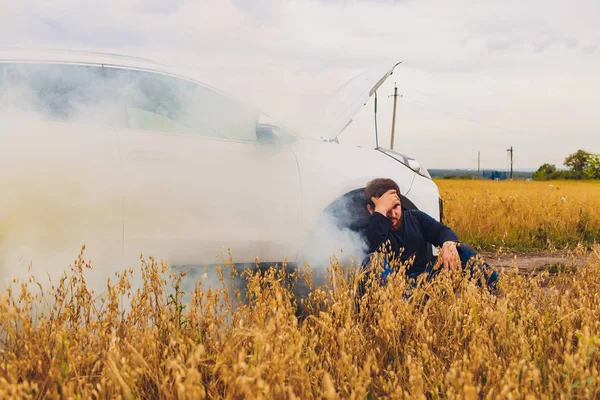 Stressed and frustrated driver pulling his hair while standing on the road next to broken car. Road trip problems and assistance concepts. smoke.