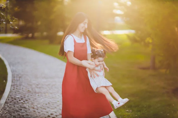 Молода мати тримає свою дочку-малятко на руках, коли вони проходять через парк в сонячний день . — стокове фото