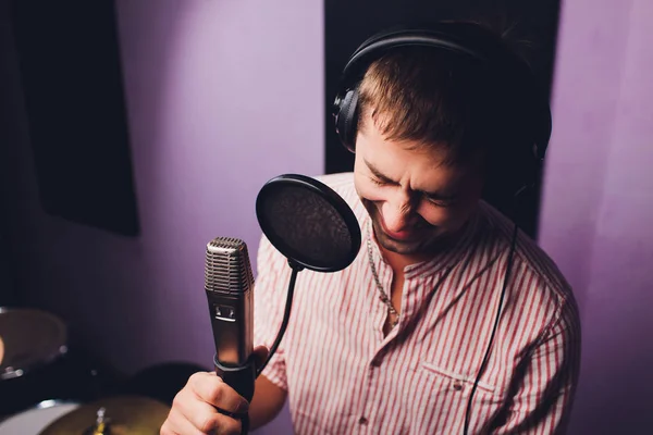 music, show business, people and voice concept - male singer with headphones and microphone singing song at sound recording studio.