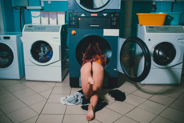 Young woman searching clothes in washing machine drum at laundromat. naked in black thong — Stock Photo, Image