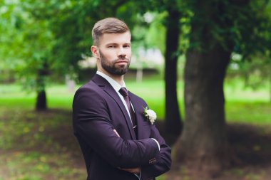 Kareli takım elbiseli yakışıklı, zarif sakallı damat ormandaki köprüde duruyor. Şık bir düğün damadı fotoğrafı. Köylü hipster portresi..