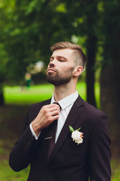 Bonito e elegante noivo barbudo em terno xadrez está em pé na ponte na floresta. Foto de noivos de casamento elegante. Retrato hipster rústico. — Fotografia de Stock