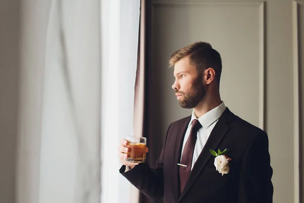 Portret van een bebaarde jongeman met retro look met een glas whisky op grijze achtergrond. — Stockfoto