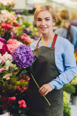 Çiçekçi olarak çalışan ve arka planda müşterisi olan bir kadın satış görevlisi. Yatay şekil, bel yukarı.