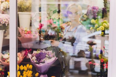 Çiçekçi olarak çalışan ve arka planda müşterisi olan bir kadın satış görevlisi. Yatay şekil, bel yukarı.