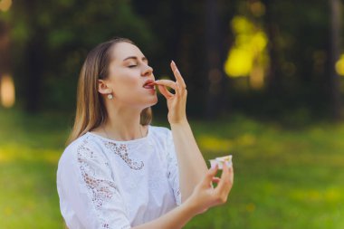 Gülümseyen kadın dışarıda öğle yemeği molası veriyor, çimlerin üzerinde oturuyor ve yemek yiyor..