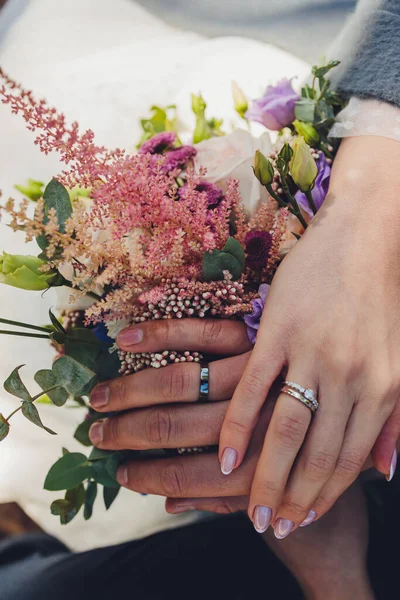 Closeup άποψη των νεόνυμφων χέρια κρατώντας πολύχρωμο γαμήλιο μπουκέτο. Νύφη και γαμπρός φοράνε βέρες. Εξωτερικό φόντο. Έννοια ημέρας γάμου. — Φωτογραφία Αρχείου