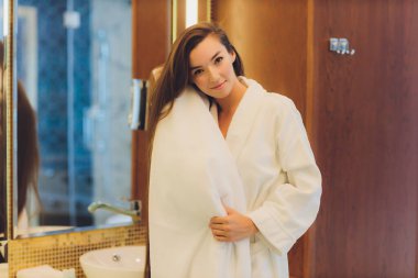 Genç bir kadın banyoda duruyor ve saçını havluyla kurutuyor. O, aynaya bakıyor. Yatay çerçeveli çekim.