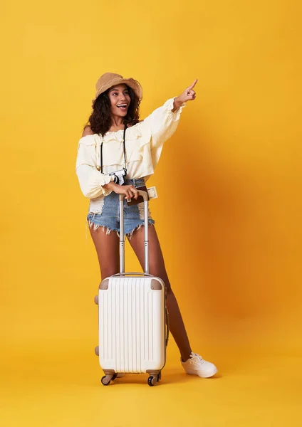 Cheerful young african woman dressed in summer clothes standing with a suitcase and pointing finger at copy space isolated over yellow background. — Stock Photo, Image