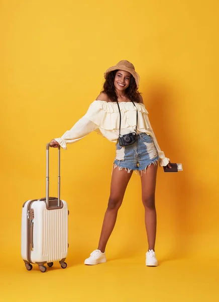 Sarı arka plan üzerinde izole bir bavul ile ayakta iken para ile pasaport tutan yazlık giysiler giymiş Neşeli genç Afrikalı kadın.