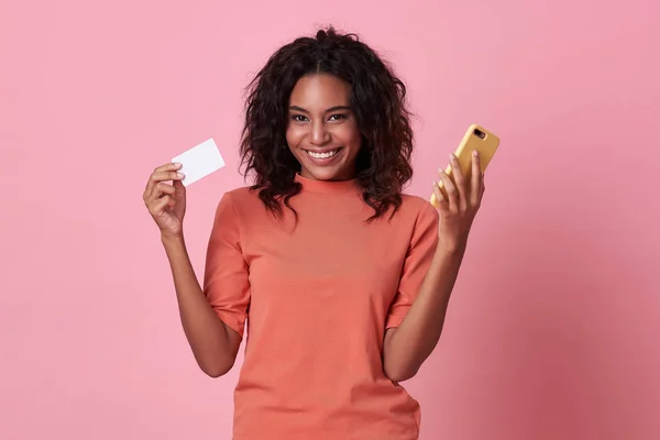 Портрет молодої африканки, що показує кредитну картку та мобільний телефон ізольовані на рожевому фоні . — стокове фото