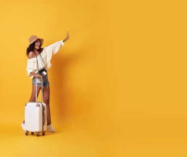 Neşeli genç afrikalı kadın yazlık giysiler giymiş bir bavul ile ayakta ve sarı arka plan üzerinde izole kopya alanda merhaba demek.