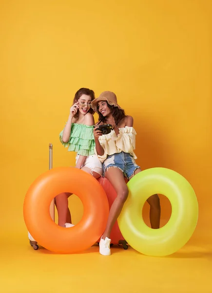 Alegre amigos mujer vestida con ropa de verano sentada en una maleta y anillo de goma aislado sobre fondo amarillo . — Foto de Stock