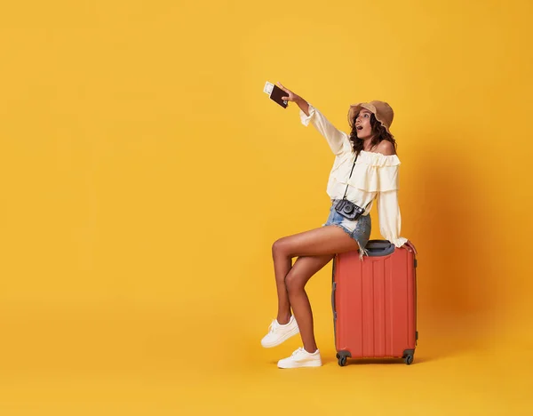 Joyeux jeune femme africaine vêtue de vêtements d'été assis sur une valise et pointant du doigt l'espace de copie isolé sur fond jaune . — Photo