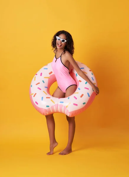 Feliz joven africana sobre fondo amarillo vestida con traje de baño sosteniendo anillo de goma playa . — Foto de Stock