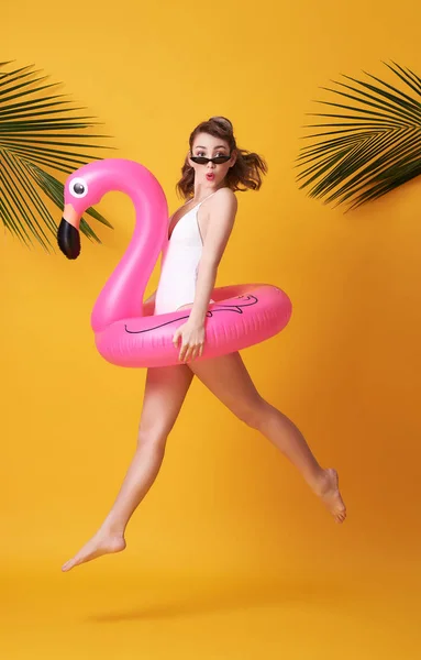 Счастливая молодая женщина прыгает на желтом фоне, одетая в купальники, держа фламинго резиновое кольцо пляжа. — стоковое фото