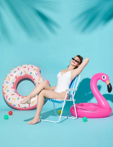 Alegre atractivo alegre deliciosas mujeres vestidas con trajes de baño agradable sentado en una silla de playa y anillo de goma sobre fondo azul brillante . — Foto de Stock