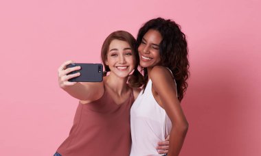 İki neşeli kız pembe arka plan üzerinde birlikte jest gösterirken selfie çekiyor.