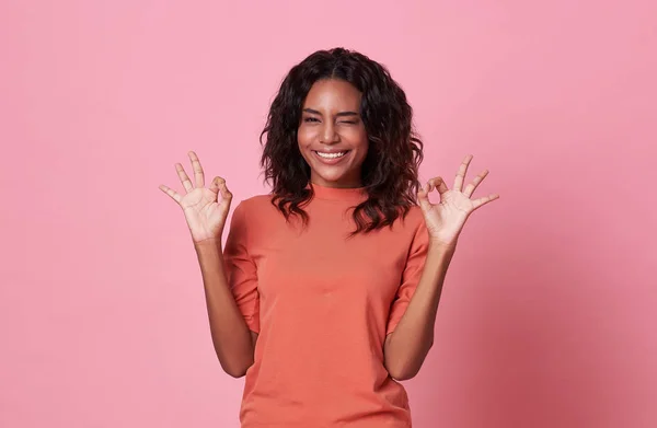 Захоплена красива африканська жінка показує добре жест, дивлячись на камеру в повсякденній помаранчевій футболці ізольовані на рожевому фоні . — стокове фото