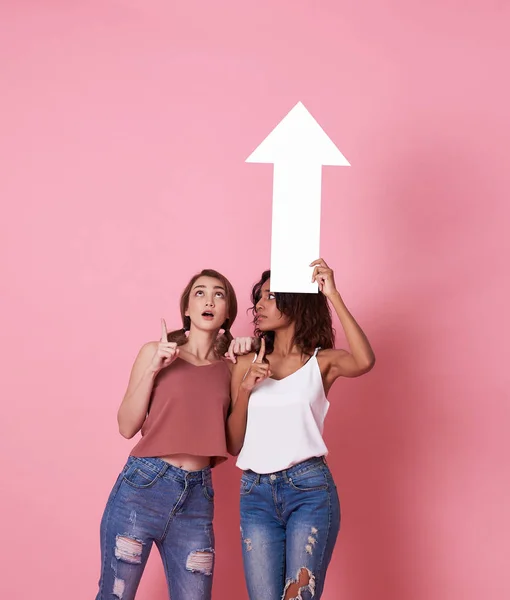 Retrato de dos jóvenes conmocionadas de pie con su flecha apuntando al espacio de copia aislado sobre el fondo de la pancarta rosa . — Foto de Stock