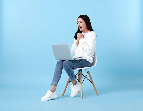 Young woman asian happy smiling in casual white cardigan with denim jeans.While her using laptop sitting on white chair isolate on bright blue background.