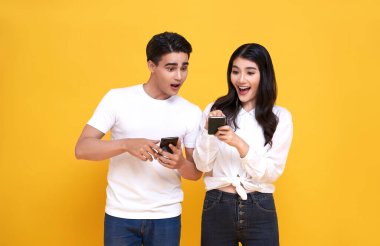İnanılmaz çekici genç Asyalı çift, sarı arka planda izole edilmiş akıllı telefonlara birlikte mesaj bakıyor..
