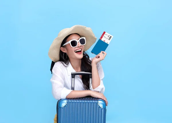 Happy young Asian tourist woman holding passport and boarding pass with baggage going to travel on holidays on blue background.