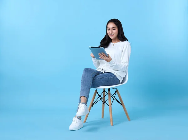 Young woman asian happy smiling in casual white cardigan with denim jeans.While her using tablet computer sitting on white chair isolate on bright blue background.
