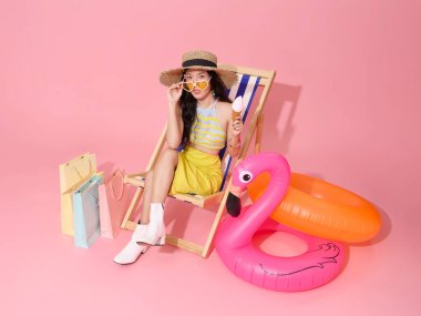 Mutlu gülümseyen Asyalı kadın yaz kıyafetleri içinde plaj sandalyesinde oturuyor yaz için alışveriş çantası satışı pembe arka planda..