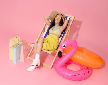 Mutlu gülümseyen Asyalı kadın yaz kıyafetleri içinde plaj sandalyesinde oturuyor yaz için alışveriş çantası satışı pembe arka planda..