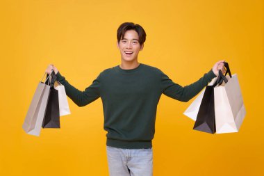Genç Asyalı alışverişçi, elinde alışveriş çantası tutarken mutlu oluyor. Sarı arka planda izole edilmiş indirim terfisi için heyecanlanıyor..