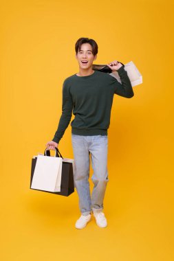 Uzun boylu Asyalı alışveriş torbası taşıyan genç adam sarı arka planda izole edilmiş indirimli promosyon için heyecanlanıyor..