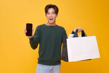 Heyecanlı Asyalı adam sarı arka planda izole edilmiş alışveriş torbalarını tutarken boş ekranlı bir akıllı telefonu gururla sergiliyor. Mobil alışveriş veya uygulama promosyonlarını göstermek için ideal.