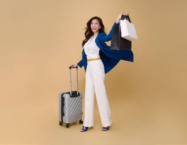 Mutlu, genç, zarif Asyalı iş kadını seyahat valizleri ve alışveriş çantası çıplak renk arka planında izole edilmiş. Turist kız neşeli bir tatil konsepti yaşıyor. 