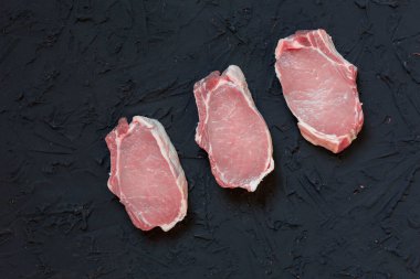 Dağılmış un, biber, sarımsak, karanfil, defne yaprağı, soğan ve otlarla kaplı koyu dokulu bir yüzeye yerleştirilmiş üç taze domuz pirzolası. Pişmemiş domuz eti pişirmeye hazır.