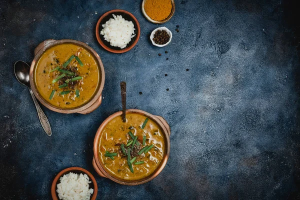 hot tasty vegetables soup with green onion in bowls and plate with rice and spices on dark blue background, close view