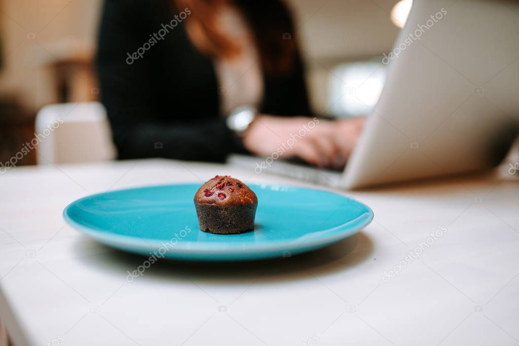 Muffin on a torquise plate, woman working on laptop on background.