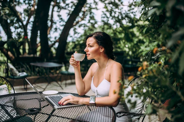 Beautiful woman drinking coffee and using laptop at garden cafe.