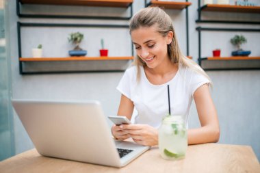 Limonata içmek ve internette gezinmek genç kadın. Telefon ve dizüstü bilgisayar kullanarak.