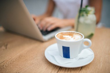 Yakın çekim görüntüsü cappuccino bir tablo. Arka planda dizüstü bilgisayar kullanan bulanık kız.
