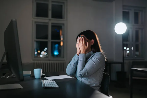 Overwhelmed businesswoman holding head in hands while working overtime at office desk.