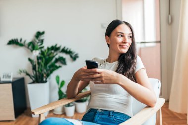 Evde akıllı telefon kullanan rahat bir genç kadın portresi. Uzağa arıyorsunuz.