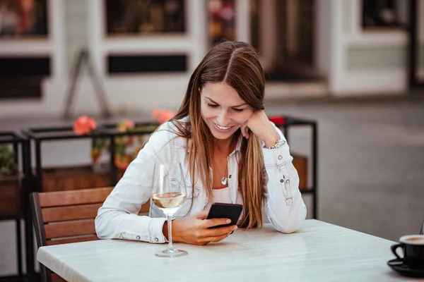 快乐的年轻女子坐在户外的餐厅里 看着智能手机 桌上的白葡萄酒杯 — 图库照片