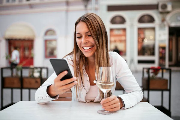 快乐的年轻女子使用智能手机 并有一杯白葡萄酒在户外餐厅 — 图库照片