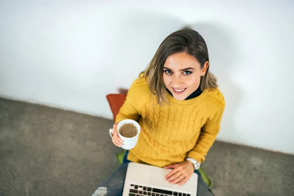 一个休闲的年轻女子的肖像 在她的腿上的笔记本电脑 拿着咖啡 看着相机 — 图库照片