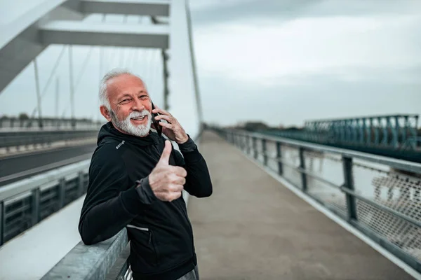 Portrait of a senior sportsman talking on the mobile phone and showing thumbs-up in the city.